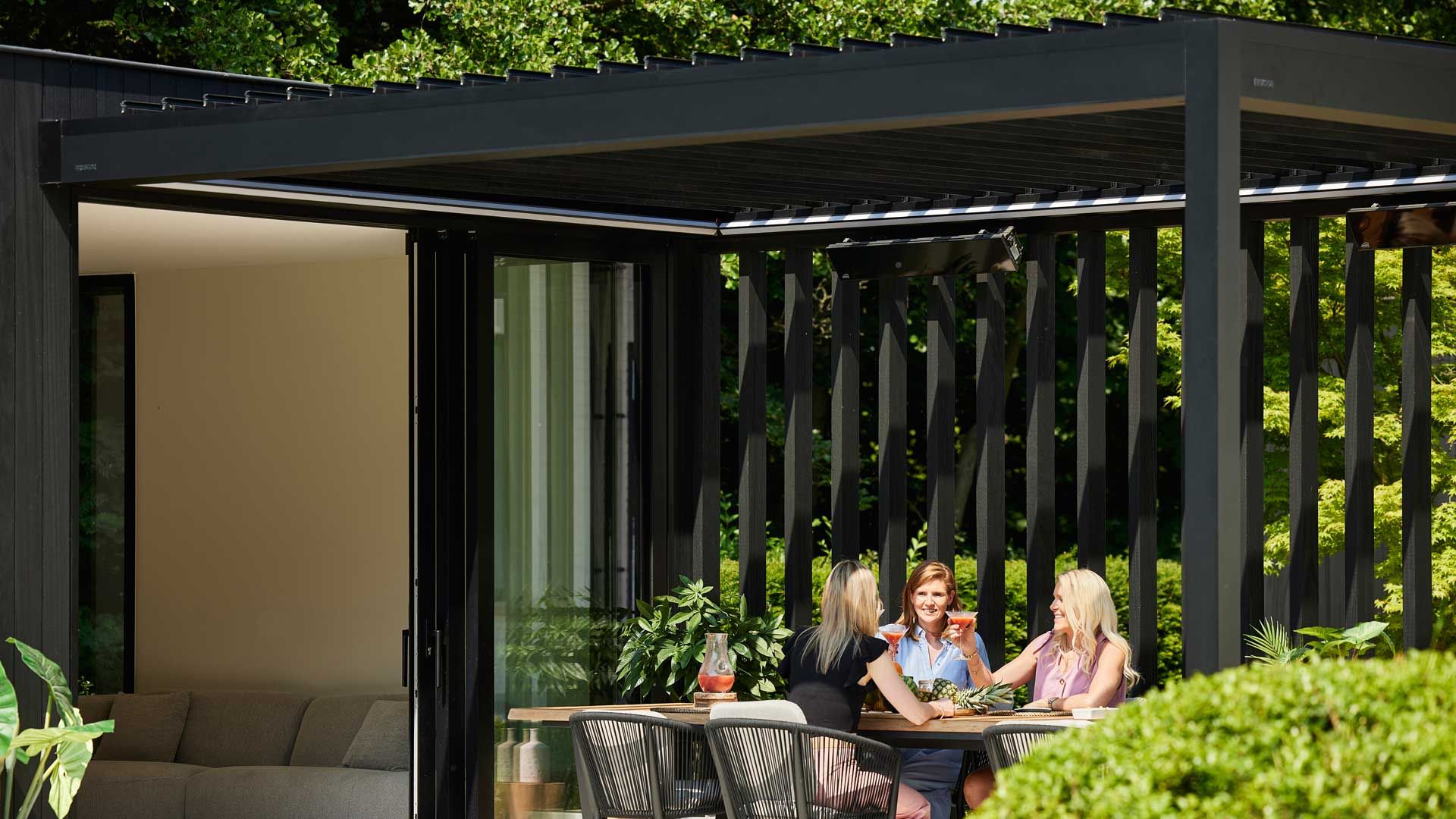 Drei Frauen sitzen mit ihren Getränken unter einer Terrassenüberdachung, im Hintergrund kann man in den Wohnzimmerbereich blicken.
