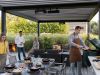 Zwei Männer und zwei Frauen stehen unter einer Terrassenüberdachung mit Grill, gedeckten Tisch und einer Sitzecke.