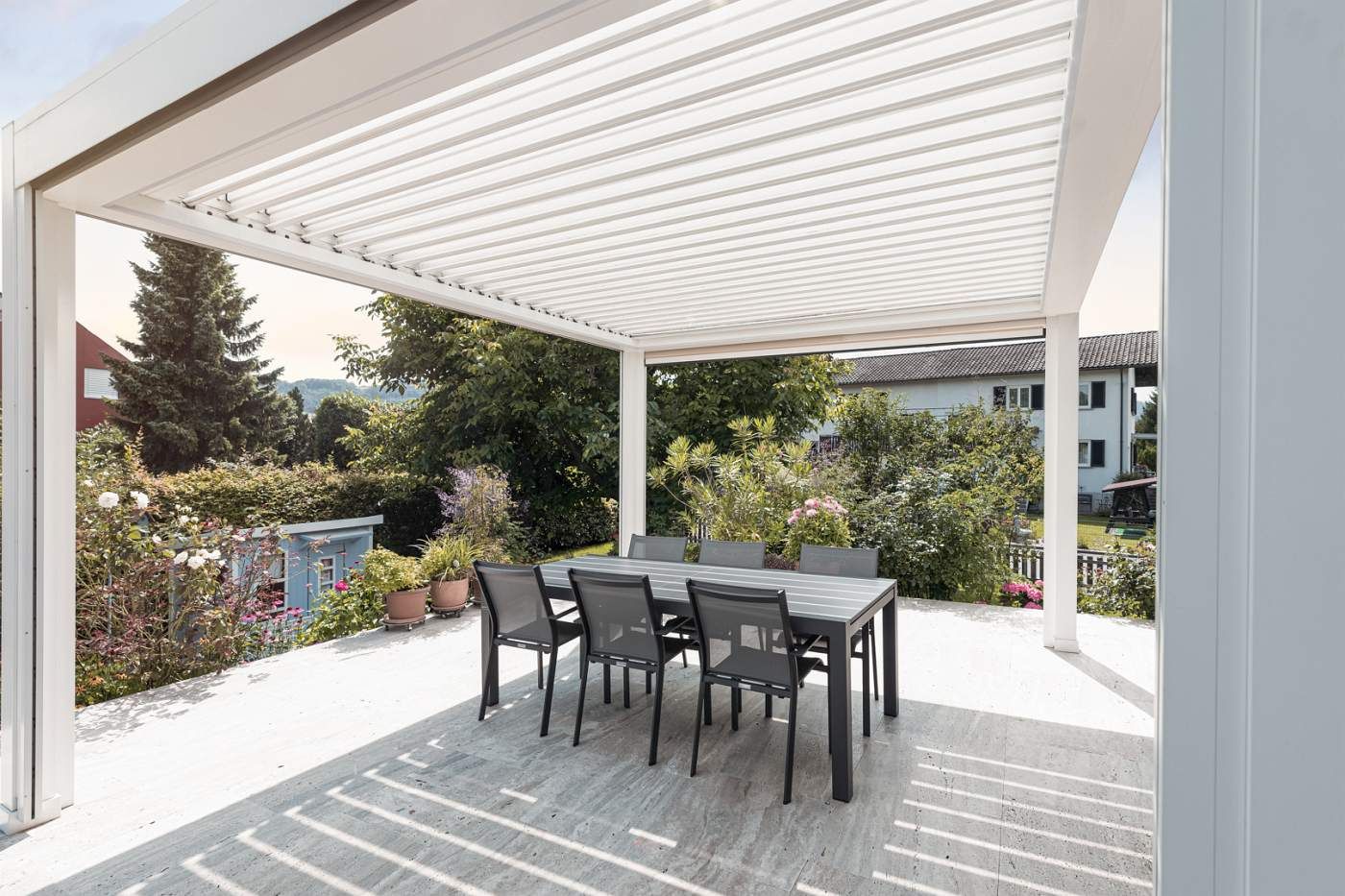 Sechs Stühle mit einem Tisch in der Mitte stehen unter einer offenen, weißen Terrassenüberdachung.