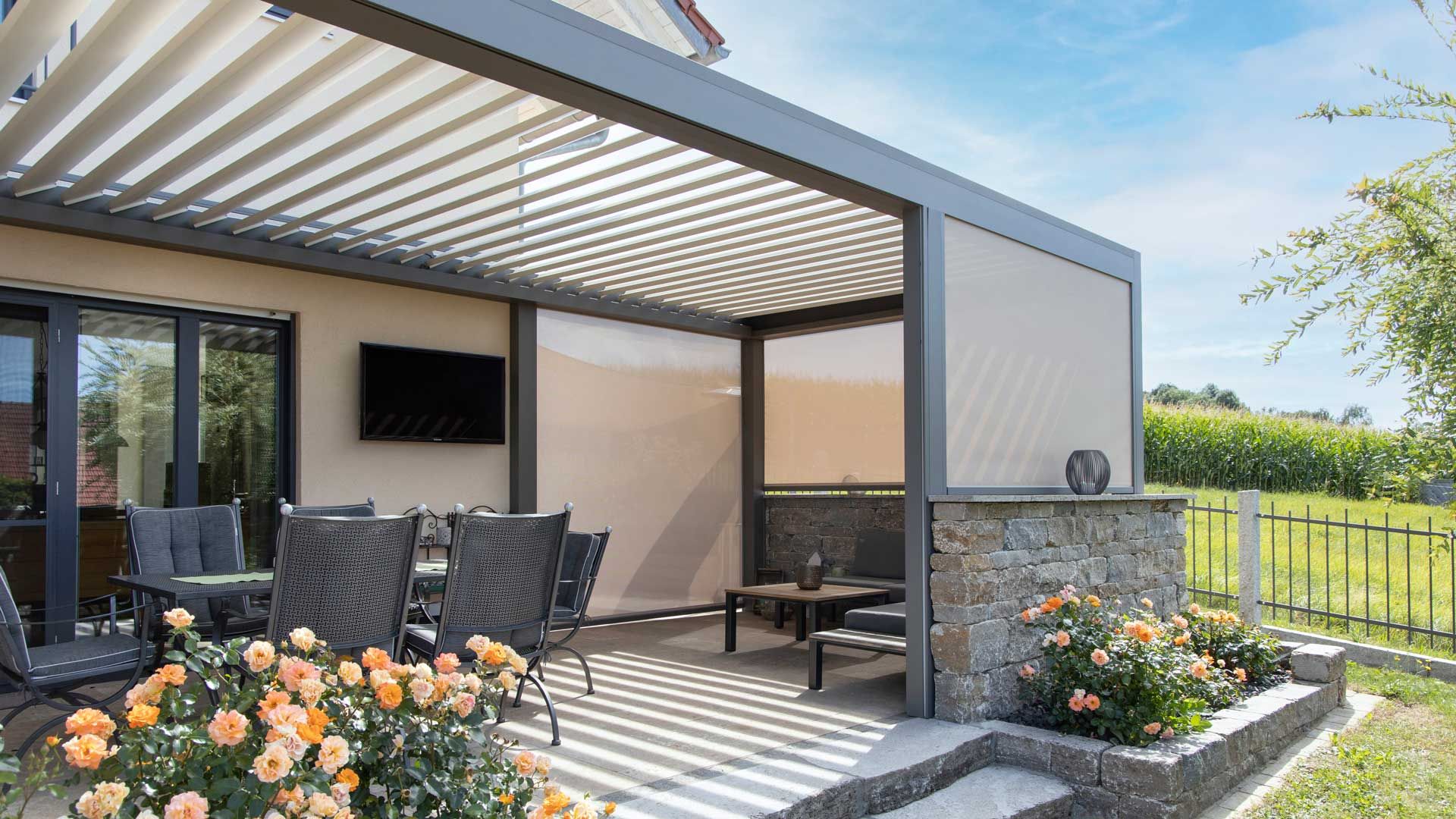Die dunkelgraue Terrassenüberdachung mit Jalousien an den Seiten und einem Maisfeld im Hintergrund.