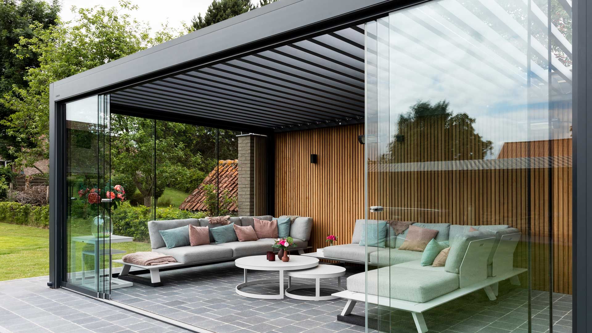 Eine Terrassenüberdachung mit einer Holzlamellen Rückwand und drei Glaselementen an den verbleibenden Seiten. Darunter zwei graue Sitzgruppen.