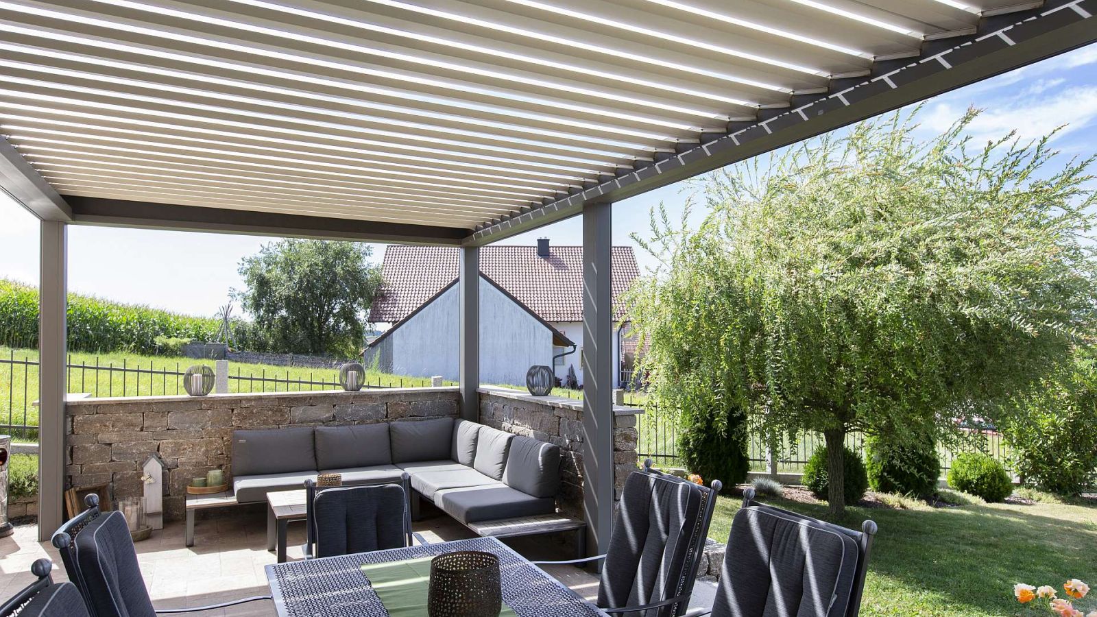 Ein Terrassendach mit Lamellen. Darunter eine Sitzgruppe und im Hintergrund der Garten.
