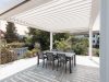 Sechs Stühle mit einem Tisch in der Mitte stehen unter einer offenen, weißen Terrassenüberdachung.