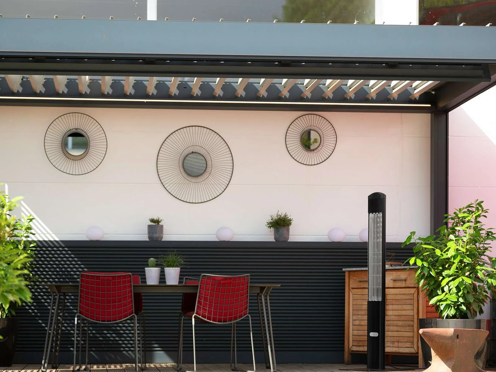 Unter einer Terrassenüberdachung steht neben einem Tisch mit vier roten Stühlen ein schwarzer Heat Tower.
