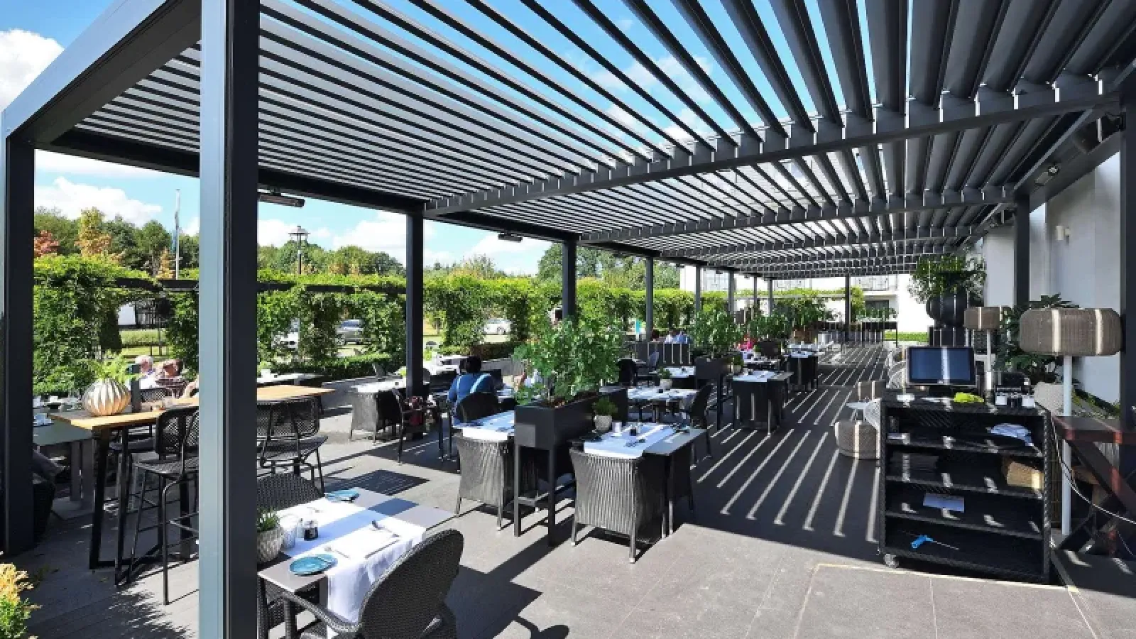 Sehr viele Tische stehen unter einen Lamellen Terrassendach, es scheint sich um eine Gastronomie zu handeln.
