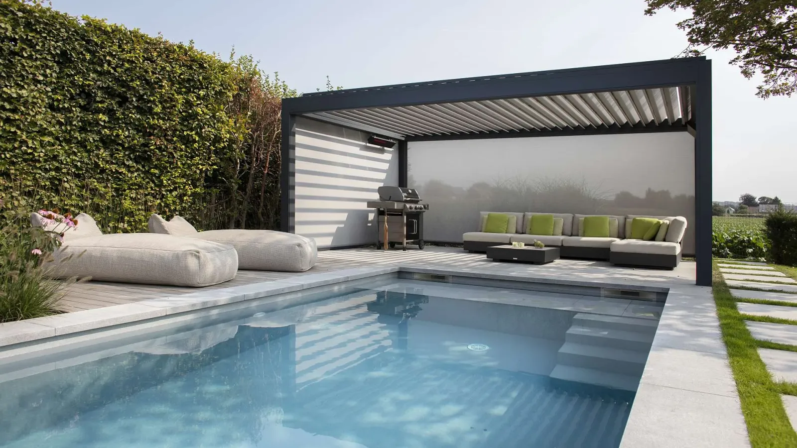 Hinter einem großen Pool befindet sich ein freistehendes Lamellendach mit Grill und Couch darunter.