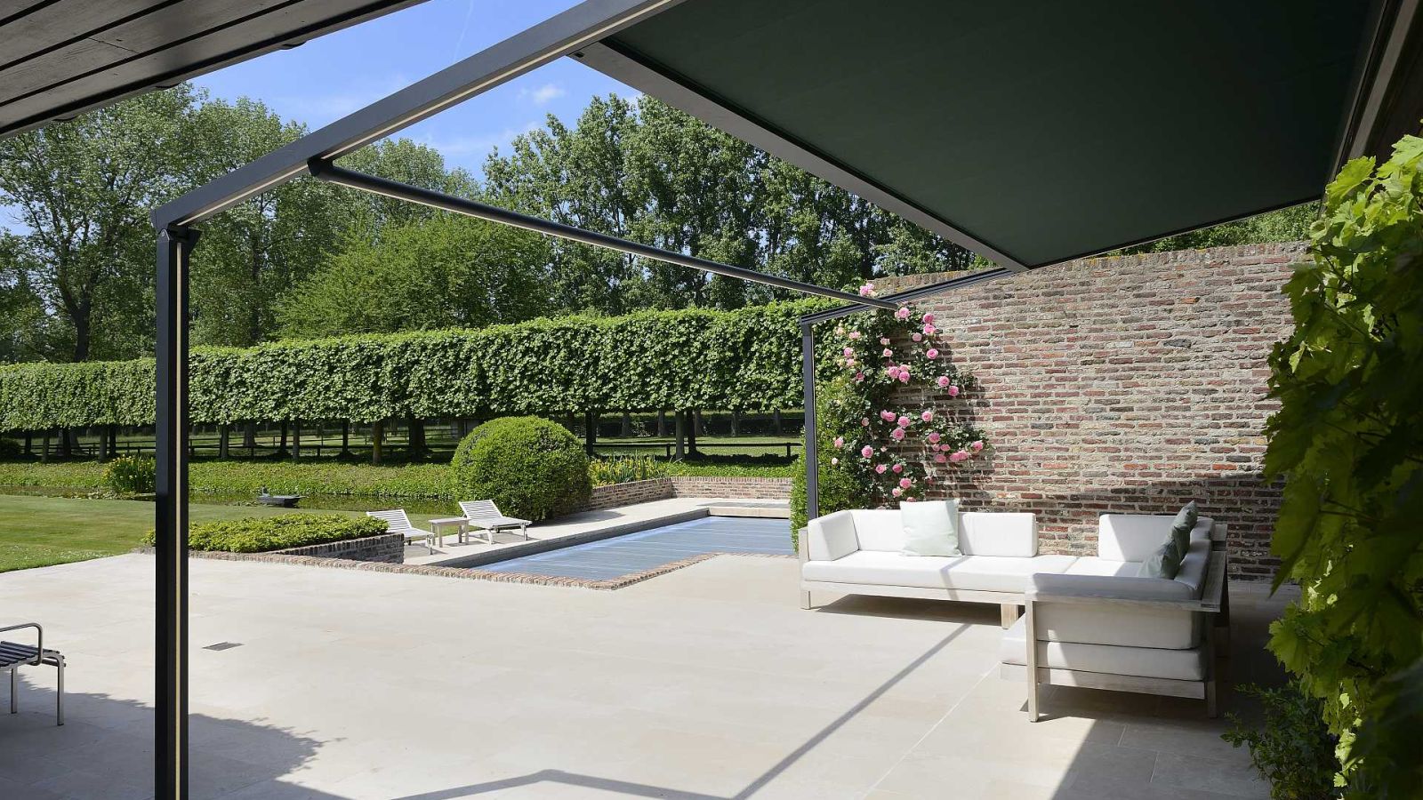Eine maßgefertigtes Terassendach Pergola mit Couch. Im Hintergrund eine Hecke und Bäume.